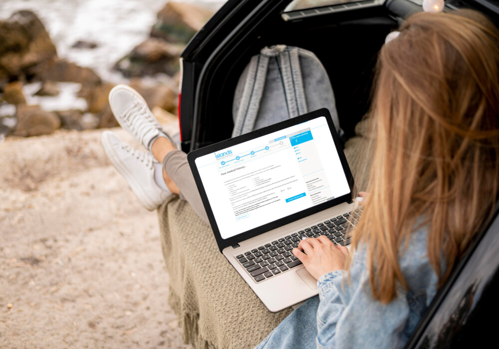 Woman relaxing in her car boot while using a laptop displaying the Islands Insurance travel insurance medical screening webpage, planning for her journey with peace of mind.