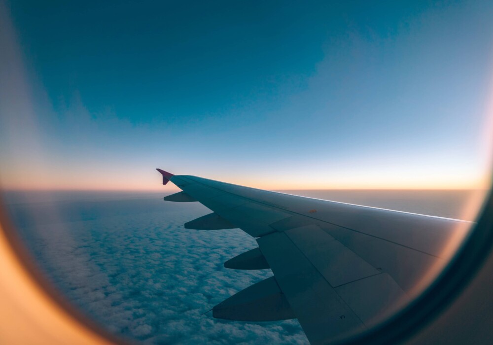 View from an airplane window during flight, showcasing a clear sky with soft gradients of blue, a sea of white clouds below, and a wingtip with a red accent against the horizon, evoking the need for Travel Insurance.